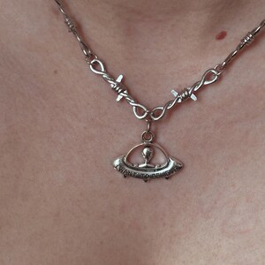 May include: Silver-toned necklace featuring a barbed wire chain and a pendant shaped like a UFO. The pendant has a small alien figure inside and the words "I WANT TO BELIEVE" are inscribed on the bottom.