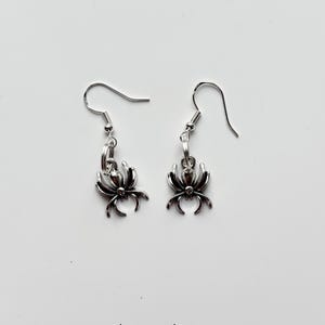 Può includere: Un paio di orecchini a forma di ragno color argento. Ogni orecchino presenta un ciondolo a ragno con otto zampe e un gancio. Lo sfondo è bianco. Il testo "Pissy Prints" è in basso.