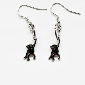 Puede incluir: Un par de pendientes de mono plateados. Cada pendiente presenta un pequeño dije de mono colgando de un alambre para la oreja en forma de anzuelo. El mono está representado con los brazos levantados, como si estuviera colgado de una rama.