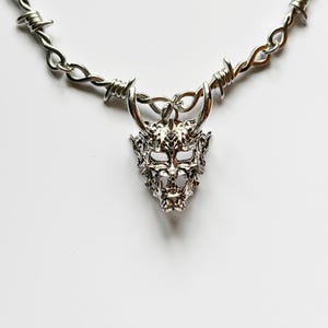May include: Silver-toned necklace featuring a detailed demon mask pendant. The necklace has a barbed wire design, adding a unique and edgy aesthetic. The pendant showcases intricate facial features, creating a bold statement piece.