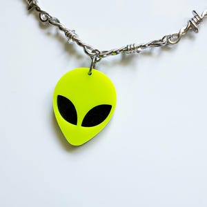 May include: A necklace featuring a silver-toned chain designed to resemble barbed wire. A bright neon yellow alien head pendant with two solid black oval-shaped eyes is attached to the center of the chain with a small silver-toned metal jump ring.