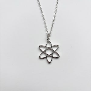 May include: A silver necklace featuring an atom-shaped pendant. The pendant is a six-pointed star design with curved lines. The chain is silver and has a delicate link pattern. The necklace is set against a plain white background.