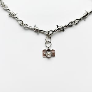 May include: Silver-toned necklace featuring a barbed wire design. A small, detailed camera charm hangs from the necklace. The camera charm has a brown and silver finish. The necklace is displayed against a white background.