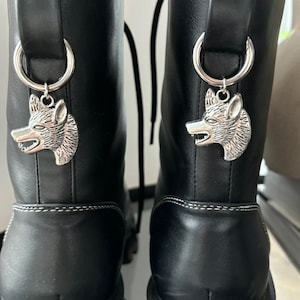 Może przedstawiać: Para czarnych skórzanych butów z srebrnymi zawieszkami w kształcie wilczej głowy przymocowanymi do sznurowadeł. Buty mają grubą platformę i białe szwy wokół górnej części.