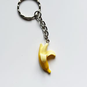 May include: A novelty banana keyring. The yellow banana has a peeled section, attached to a silver chain and keyring. The banana is a bright yellow with a lighter yellow peel. A fun, quirky accessory.