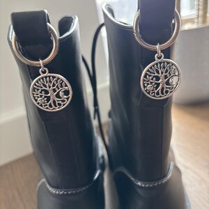 Può includere: Due stivali in pelle nera con ciondoli color argento. Ogni ciondolo è un pendente circolare con un disegno ad albero e una luna crescente. I ciondoli sono attaccati agli stivali con anelli d'argento.