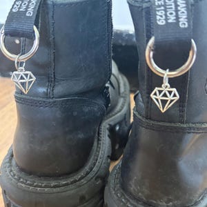 Op de afbeelding: Close-up van twee zwarte leren laarzen met dikke zolen. Elke laars heeft een zilveren bedel in de vorm van een diamant die aan een zilveren ring hangt. De laarzen hebben de tekst "REMAKING TRADITION SINCE 1929" op de treklip.