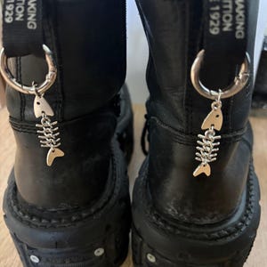May include: Pair of black leather platform boots with silver fishbone charms hanging from the top. The boots have a chunky sole and a strap with the text "OEMAKING TRADITION SINCE 1929".