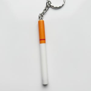 May include: A novelty keychain shaped like a cigarette. The keychain features a white, cylindrical body with an orange tip and a red band. It is attached to a silver chain and keyring.