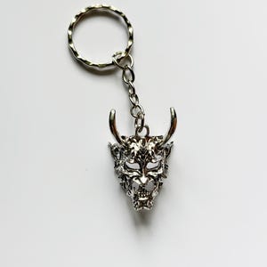May include: Silver-colored keychain featuring a detailed oni mask pendant. The mask has intricate facial features, horns, and a textured surface. The keychain includes a split ring and a chain for attaching keys or accessories.