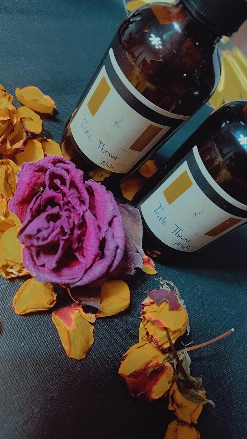 May include: Two brown glass bottles with gold labels that say "Triple Threat MLS". The bottles are sitting on a dark blue surface with dried rose petals.