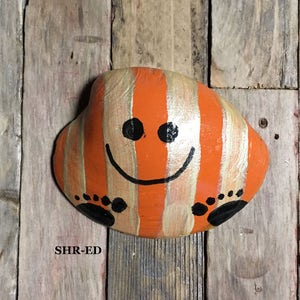 May include: A hand-painted rock with orange and gold stripes, a smiling face, and black footprints. The rock is on a wooden background.