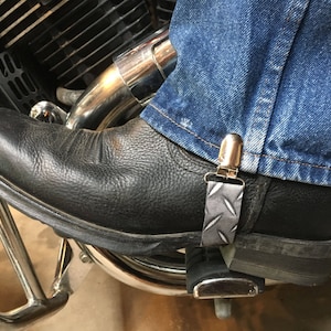 May include: Close-up of a black leather motorcycle boot with a silver and gray patterned boot strap. The boot is paired with blue denim jeans. The boot is on a motorcycle.