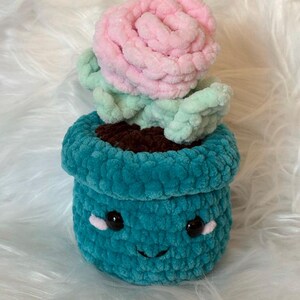 Puede incluir: Una planta suculenta de crochet en una maceta de color turquesa con una cara sonriente. La suculenta tiene hojas rosas y verdes.