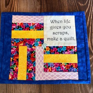 May include: A quilted placemat with a blue border and a patchwork design. The patchwork features floral fabric, yellow stripes, and a white square with the text "When life gives you scraps, make a quilt."