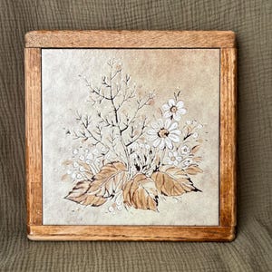 May include: A square ceramic tile set within a natural wood frame. The tile features a hand-painted illustration of a floral bouquet with white daisy-like flowers, brown leaves, and delicate stems against a light beige, textured background.