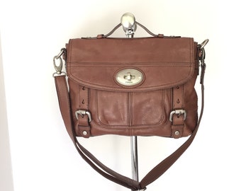 May include: A brown leather Fossil satchel bag with a silver buckle and a silver logo plate. The bag has a long adjustable strap and a top flap closure.