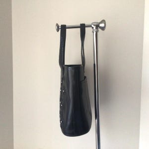 May include: A black leather tote bag with a silver buckle and a decorative design. The bag is hanging on a silver stand.