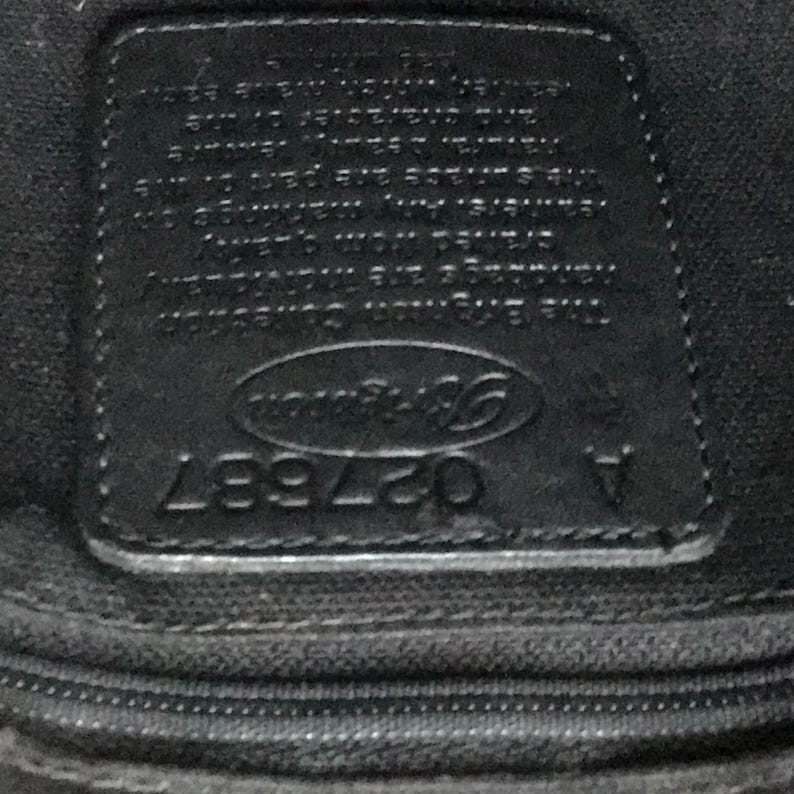 May include: Close-up of a black leather tag with the text "Coach" in a white oval and the number "027997" below it.