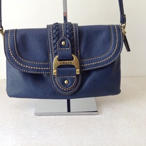 May include: A navy blue crossbody bag with a flap closure and a braided accent. The bag features gold-colored hardware, including a buckle with the brand name "CHAPS". The stitching is a contrasting tan color.