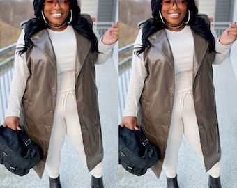 May include: A long brown faux leather vest with a collared neckline and button closure. The vest is worn over a white long-sleeved shirt and cream-colored sweatpants. The person is wearing black ankle boots and carrying a black bag.