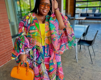 May include: A woman wearing a colorful, patterned kimono-style garment with a yellow top and orange platform sandals. She is holding an orange handbag and wearing orange-tinted sunglasses. The kimono features a vibrant mix of pink, green, and blue floral designs.