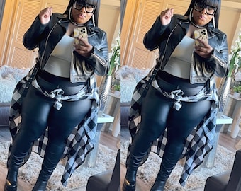 May include: A woman wearing a black leather jacket, a white tank top, black leather pants, and a black and white plaid shirt tied around her waist. She is wearing black boots and has her hair pulled back in a ponytail. She is holding a cell phone in her hand and is looking at the camera.