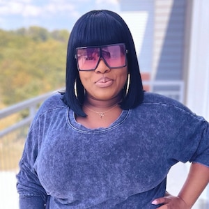 May include: A woman with black hair wearing a blue, textured, long-sleeved shirt and large, black square sunglasses with pink lenses.