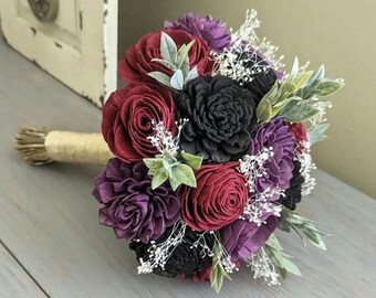 May include: A burgundy, purple, and black sola wood flower bouquet with greenery and white baby's breath.