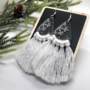 May include: A pair of dangle earrings featuring clear, teardrop-shaped acrylic charms etched with white snowflake patterns. Each charm is attached to a long, thick white cotton tassel with a knotted top, suspended from silver-toned metal hooks.
