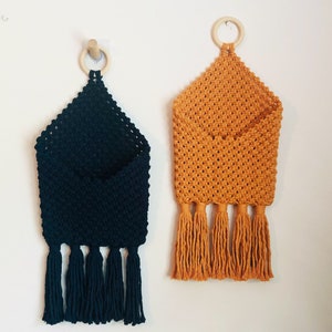 May include: Two macrame wall pockets, one in dark navy blue and one in mustard yellow. Each pocket features a knotted cord pattern, a wooden ring at the top for hanging, and four long, dangling tassels at the bottom edge of the fabric structure.