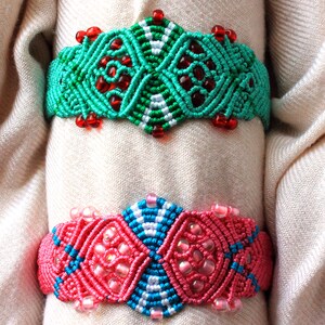 May include: Two macrame bracelets. The top bracelet is turquoise with red beads and white accents. The bottom bracelet is pink with blue and white accents and pink beads. Both bracelets are displayed on a beige fabric.