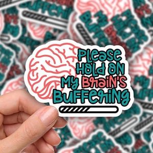 May include: A hand holding a white sticker with a brain graphic and the text "Please Hold On My Brain's Buffering". The sticker has a pink brain design and teal and red text. The background is a collection of similar stickers.