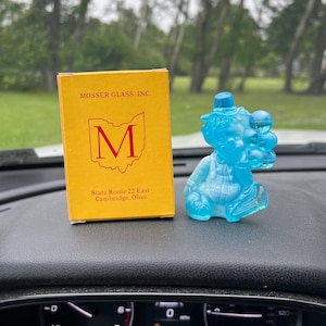 May include: A bright blue glass clown figurine holding a cluster of balloons, positioned next to a yellow cardboard box labeled MOSSER GLASS, INC. with a red M logo and the text State Route 22 East, Cambridge, Ohio printed on the front surface.