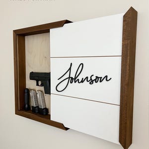 May include: A walnut-framed, white shiplap-faced portrait-style display case. The case features a black handgun, two magazines, and a flashlight. The name "Johnson" is displayed on the white face.