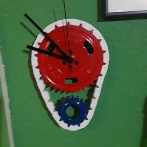 May include: A red, white, and blue clock made from three interlocking gears. The clock face is red with black hands and white numbers. The gears are painted white, blue, and red.