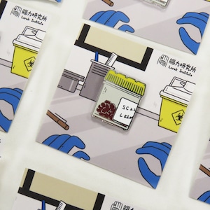 May include: A white enamel pin with a red and yellow illustration of a small jar with a label that reads "SC 22 L KIM". The jar contains red, round objects. The pin is on a white card with a blue glove and a yellow biohazard container.