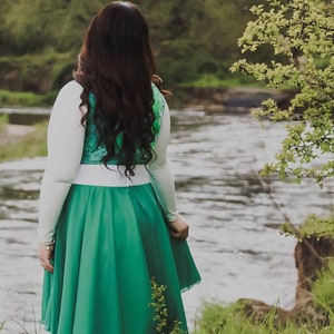 Puede incluir: Una mujer con una camisa blanca de manga larga, un chaleco verde y blanco y una falda verde se encuentra junto a un r&iacute;o. La falda es una falda circular con una cintura blanca. La mujer tiene el pelo largo y oscuro.