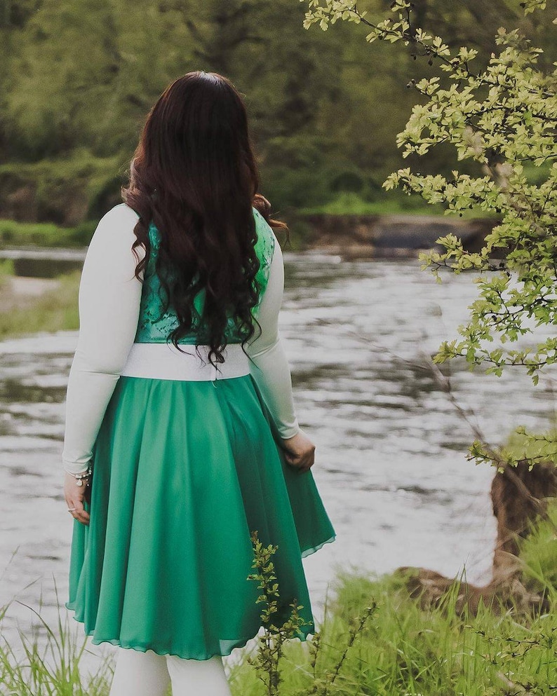 Puede incluir: Una mujer con una camisa blanca de manga larga, un chaleco verde y blanco y una falda verde se encuentra junto a un r&iacute;o. La falda es una falda circular con una cintura blanca. La mujer tiene el pelo largo y oscuro.