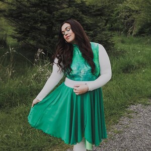 Puede incluir: Una mujer con una camisa blanca de manga larga con un corpi&ntilde;o estampado en verde y azul y una falda verde. Est&aacute; de pie en un camino con hierba verde a ambos lados.