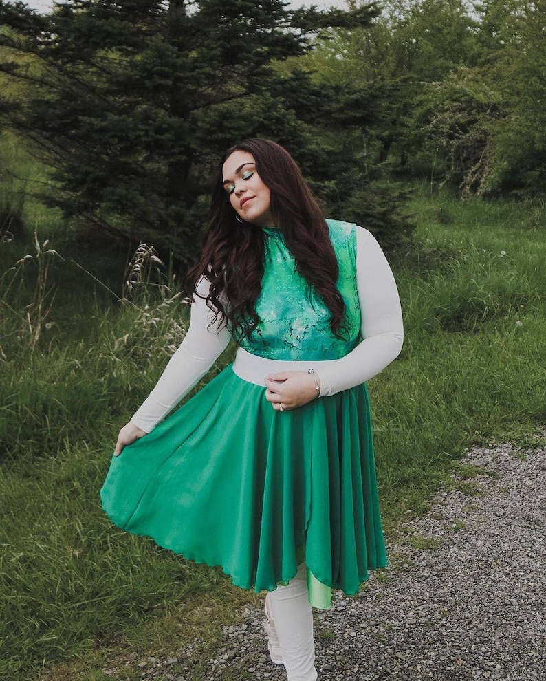 Puede incluir: Una mujer con una camisa blanca de manga larga con un corpi&ntilde;o estampado en verde y azul y una falda verde. Est&aacute; de pie en un camino con hierba verde a ambos lados.