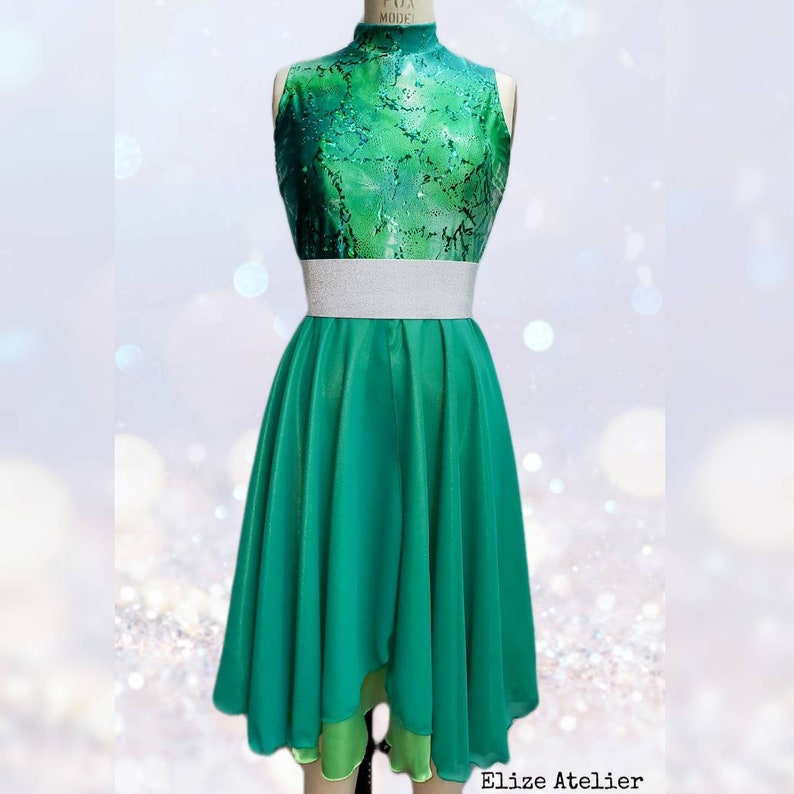 Puede incluir: Un top de baile verde sin mangas con un cintur&oacute;n plateado y una falda verde a juego con un fondo en capas. La falda es de l&iacute;nea A y tiene una cintura alta. El top tiene cuello alto y un corpi&ntilde;o ajustado. La tela es brillante y tiene un patr&oacute;n texturizado.  Elize Atelier est&aacute; impreso en la parte inferior de la imagen.