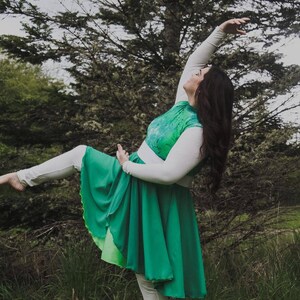 Puede incluir: Una mujer con una camisa blanca de manga larga con un corpi&ntilde;o estampado en verde y blanco y una falda verde. Est&aacute; posando con la pierna extendida y el brazo levantado por encima de la cabeza.