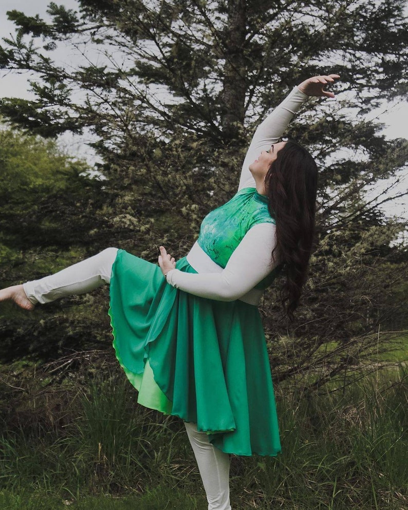 Puede incluir: Una mujer con una camisa blanca de manga larga con un corpi&ntilde;o estampado en verde y blanco y una falda verde. Est&aacute; posando con la pierna extendida y el brazo levantado por encima de la cabeza.