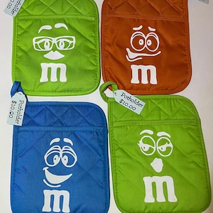 Puede incluir: Cuatro agarraderas acolchadas con diferentes caras de M&M's. Cada agarradera cuesta 10,00 €. Las caras incluyen un par de gafas, una cara sorprendida, una cara sonriente y una cara guiñando un ojo.