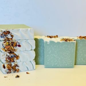 以下が含まれることがあります： 薄い緑色で白い渦巻き模様の、手作り石鹸が4つ。石鹸には、乾燥した花とハーブが埋め込まれています。