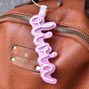 May include: A pink keychain with the word "love" in a cursive font, accented with white trim, hangs from a silver ring. The keychain is attached to a brown leather backpack with gold zippers and hardware.