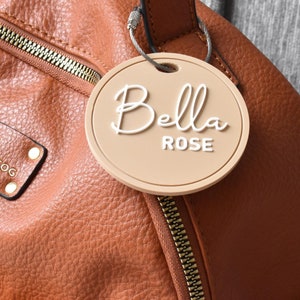 May include: A brown leather backpack with a gold zipper and a beige round tag that says "Bella Rose" in white cursive lettering.