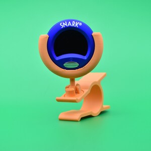 Può includere: Un accordatore per chitarra a clip blu e arancione con la scritta "SNARK" stampata sul davanti.