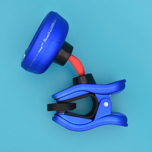 Pode incluir: Um afinador de guitarra azul com clip e pescoço flexível vermelho. O afinador tem um visor circular e um clip para prender a um instrumento.